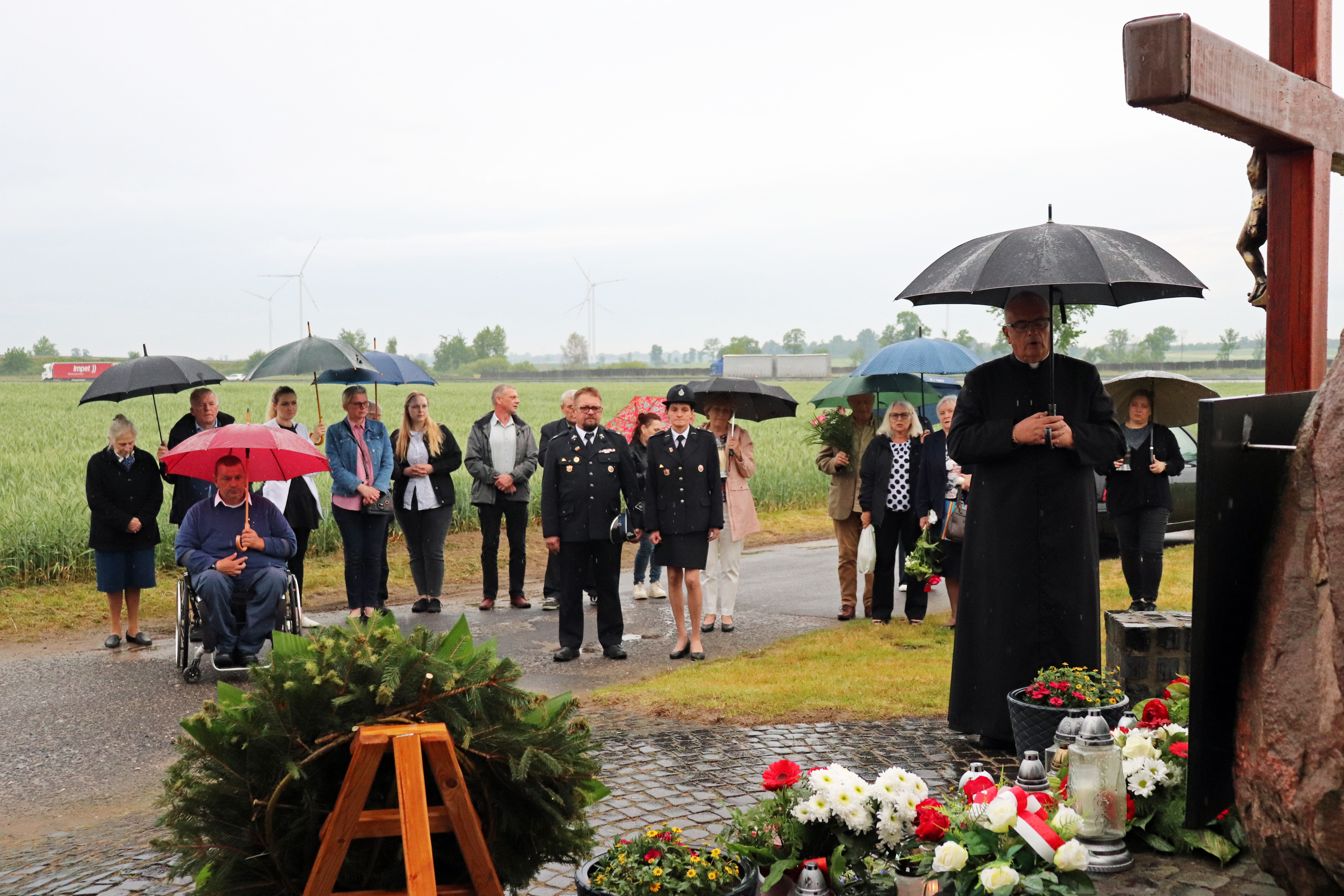 Ksiądz w trakcie modlitwy przy pomniku. W tle grupa osób, wszyscy trzymają parasole.